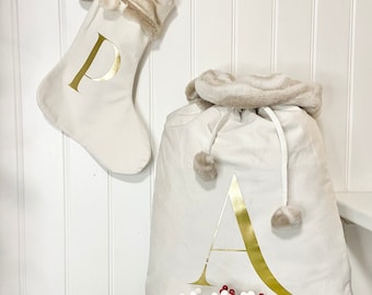 May include: Two white Christmas stockings with faux fur trim. The stocking on the left has a gold letter "P" on it. The stocking on the right is a sack with a gold letter "A" on it. Both stockings have white pom-poms.