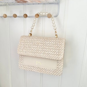 May include: A white woven straw handbag with a white leather flap and gold hardware. The handbag has a pearl handle and the initials "PS" are embossed on the flap.