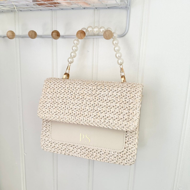 May include: A white woven straw handbag with a white leather flap and gold hardware. The handbag has a pearl handle and the initials "PS" are embossed on the flap.