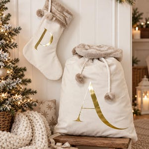May include: A cream-colored fabric Christmas stocking with a gold foil letter P and a faux fur cuff with pom-poms, paired with a matching large drawstring gift sack featuring a gold foil letter A and a plush faux fur top trim with pom-poms.