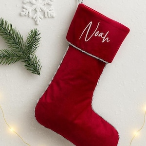 May include: A deep red velvet Christmas stocking featuring a white embroidered name Noah on the cuff. The stocking is trimmed with a black and white twisted cord edge and includes a matching loop at the top for hanging purposes.