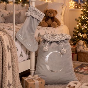 May include: A gray velvet Christmas stocking with the name HANNAH embroidered in white and a gray velvet Santa sack featuring a large silver letter C, both trimmed with a white and gray leopard print faux fur cuff and matching pom-poms.