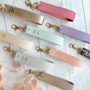 May include: Assortment of colorful leather keychains with gold metal rings and personalized initials. The keychains are displayed on a light wood surface.