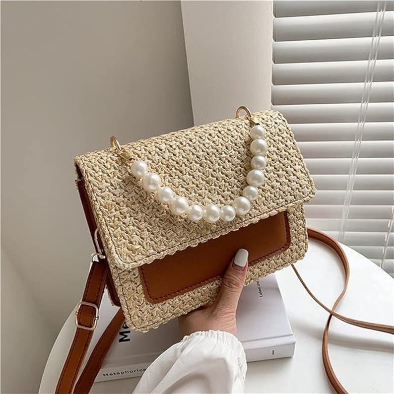 May include: A straw handbag with a brown leather strap and a pearl chain detail. The bag has a brown leather flap with a woven straw design.