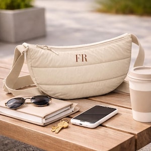 May include: A beige crescent-shaped quilted crossbody bag featuring the letters FR embroidered in a dark brown thread on the front. The bag has a wide adjustable strap and a top zipper closure, presenting a soft, padded texture throughout.