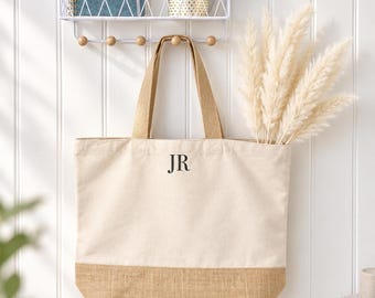 May include: A cream-colored canvas tote bag with tan burlap trim at the bottom and two matching canvas handles. The front of the bag features the black letters JR centered near the top. The bag is hanging on a hook against a white paneled wall.
