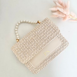 May include: A small, woven straw handbag with a pearl handle and a cream-colored flap with the initials "PS" in gold lettering.