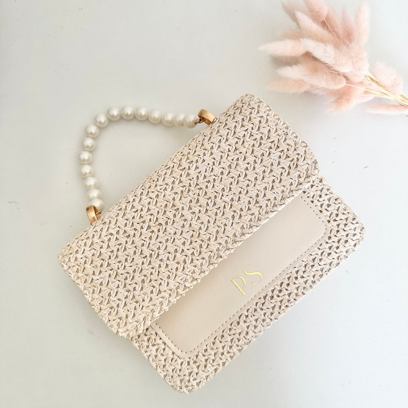May include: A small, woven straw handbag with a pearl handle and a cream-colored flap with the initials "PS" in gold lettering.