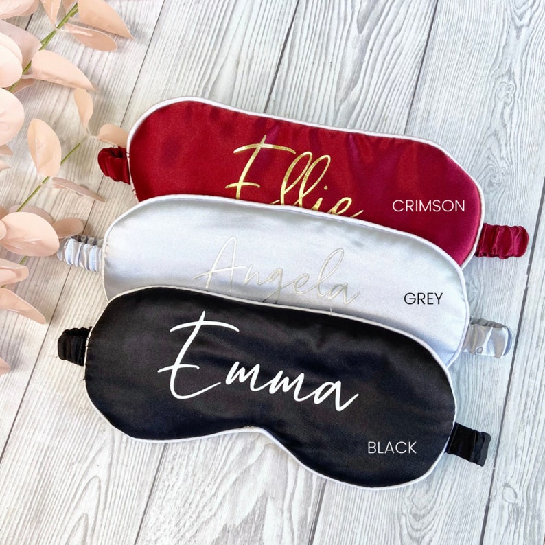 May include: Three personalized sleep masks in crimson, grey, and black with white piping. The masks are made of satin and have the names "Ellie", "Angela", and "Emma" embroidered in gold thread.
