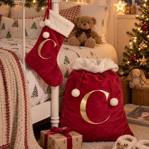 May include: A red velvet Christmas stocking and a matching large drawstring gift sack, both featuring a gold letter C. The stocking has a white faux fur cuff and pom-poms, while the sack has a white faux fur top and drawstring pom-poms.