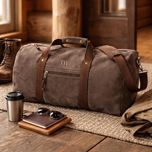 May include: A brown canvas duffel bag with brown leather accents, a zippered front pocket, and a shoulder strap. The initials "TH" are embroidered on the front. Sunglasses, a notebook, and a travel mug are next to the bag. The bag is approximately 20 inches long.