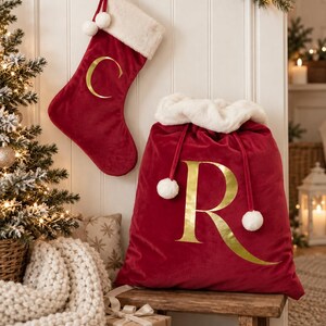 May include: A deep red velvet Christmas stocking with a white faux fur cuff and a gold letter C, paired with a matching large red velvet Santa sack featuring a white faux fur trim, gold letter R, and white pom-pom drawstring ties.