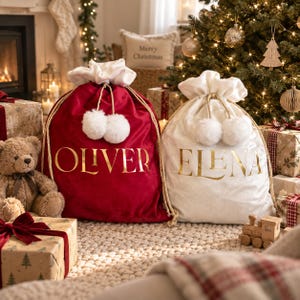 May include: Two large drawstring gift sacks, one in deep red velvet and one in white velvet, each featuring gold metallic lettering reading OLIVER and ELENA respectively. Both sacks have white faux fur trim at the top and white pom-pom accents.