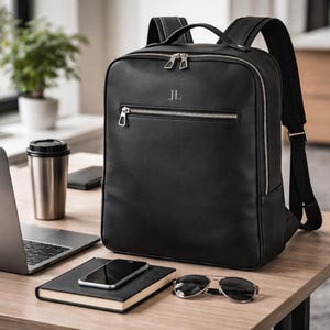 May include: Black leather backpack with silver zippers and the initials "JL" embossed on the front. The backpack has a top handle and adjustable shoulder straps. A laptop, phone, sunglasses, and a notebook are on the table.