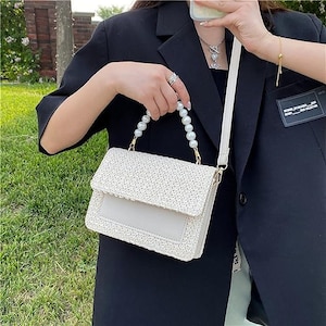 May include: A white woven handbag with a pearl strap. The bag has a flap closure and a small pocket on the front.