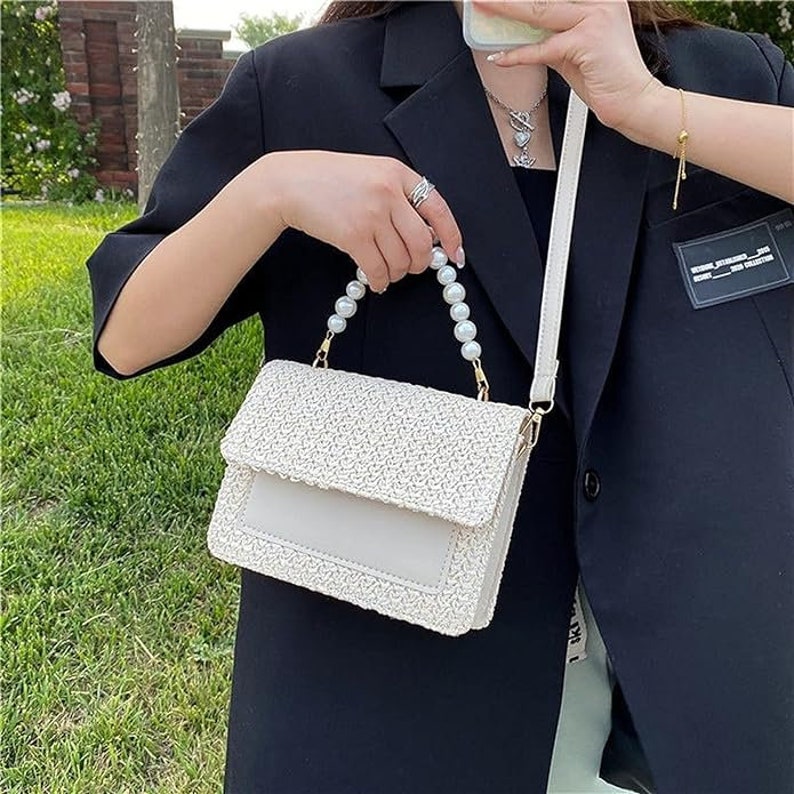 May include: A white woven handbag with a pearl strap. The bag has a flap closure and a small pocket on the front.