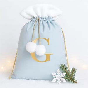 May include: A pale blue Christmas gift bag with a white, fluffy top and gold drawstring. The bag has a gold letter "G" and two white pom-poms. A white snowflake and a sprig of evergreen are placed near the bottom of the bag.