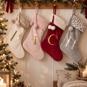 May include: Four hanging Christmas stockings in cream, light pink, deep red, and gray. Each stocking features a plush cuff, two pom-poms, and a large metallic letter: P, A, C, and H. The cream and pink stockings have gold letters, the red has gold, and the gray has silver.