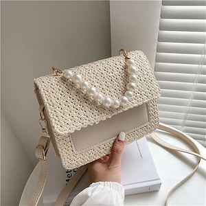 May include: A beige woven straw handbag with a pearl chain strap. The bag has a flap closure and a beige leather bottom.