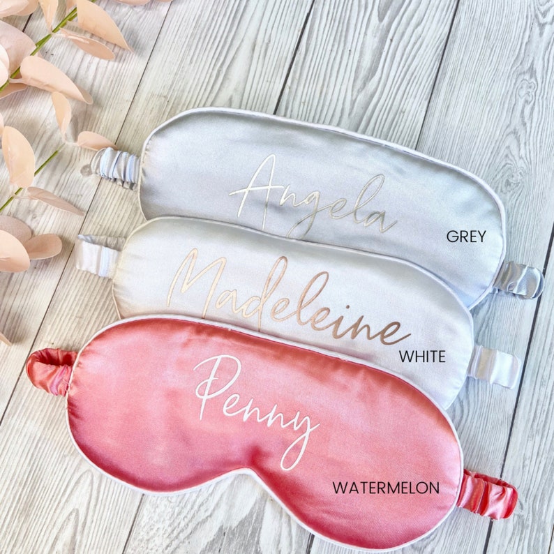 May include: Three satin sleep masks in different colors: grey, white, and watermelon. Each mask has a different name embroidered on it in gold lettering: Angela, Madeleine, and Penny.