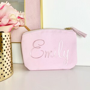 May include: A pink canvas zippered pouch with the name "Emily" embroidered in white cursive lettering.