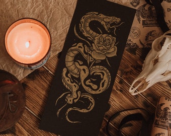 Könnte beinhalten: Eine schwarze Karte mit einer goldenen Schlangen- und Rosenillustration. Die Schlange ist um den Rosenstängel gewickelt und ihr Kopf ist erhoben, mit offenem Maul. Die Rose ist in voller Blüte und hat ein detailliertes Design.