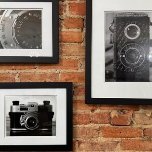 K&ouml;nnte beinhalten: Drei gerahmte Schwarz-Wei&szlig;-Fotografien von Vintage-Kameras h&auml;ngen an einer roten Backsteinmauer. Die Fotografien zeigen detaillierte Nahaufnahmen von Kameraobjektiven und -geh&auml;usen und erfassen die Essenz klassischer Fotoausr&uuml;stung.