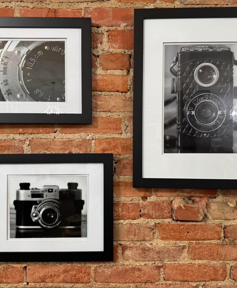 K&ouml;nnte beinhalten: Drei gerahmte Schwarz-Wei&szlig;-Fotografien von Vintage-Kameras h&auml;ngen an einer roten Backsteinmauer. Die Fotografien zeigen detaillierte Nahaufnahmen von Kameraobjektiven und -geh&auml;usen und erfassen die Essenz klassischer Fotoausr&uuml;stung.