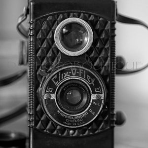 K&ouml;nnte beinhalten: Schwarz-Wei&szlig;-Vintage-Clix-V-Flex-Kamera. Die Kamera hat ein Rautenmuster auf der Vorderseite und einen Lederriemen. Das Objektiv ist mit "Maestro Lens" und "Metropolitan Industries Chicago Made in U.S.A." beschriftet.