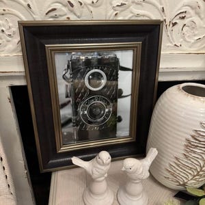 K&ouml;nnte beinhalten: Ein gerahmtes schwarz-goldenes Bild einer Vintage-Kamera. Zwei wei&szlig;e Keramikvogel-Figuren stehen vor dem Rahmen. Rechts vom Rahmen befindet sich eine wei&szlig;e Vase mit Blattmuster.
