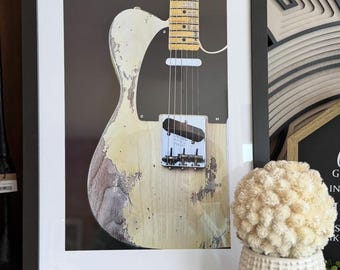 Może przedstawiać: Oprawiony wydruk zabytkowej gitary elektrycznej z postarzanym, kremowym korpusem. Gitara ma czarny pickguard, chromowany osprzęt i klonową szyjkę. Obraz jest w czarnej ramie, odpowiedni do dekoracji ścian.