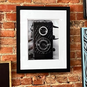 K&ouml;nnte beinhalten: Schwarz-Wei&szlig;-Fotografie einer Vintage-Kamera, gerahmt in einem schwarzen rechteckigen Rahmen mit wei&szlig;em Passepartout. Die Kamera hat zwei Objektive und ist an einer Backsteinmauer befestigt.