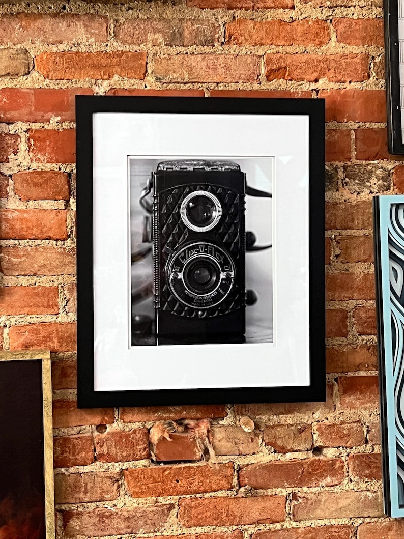K&ouml;nnte beinhalten: Schwarz-Wei&szlig;-Fotografie einer Vintage-Kamera, gerahmt in einem schwarzen rechteckigen Rahmen mit wei&szlig;em Passepartout. Die Kamera hat zwei Objektive und ist an einer Backsteinmauer befestigt.
