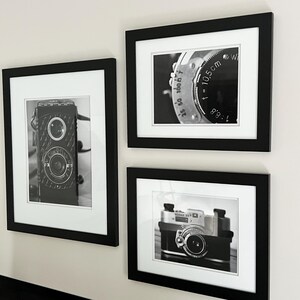 K&ouml;nnte beinhalten: Drei gerahmte Schwarz-Wei&szlig;-Fotografien von Vintage-Kameras und Kamerateilen. Die Bilder werden in schwarzen Rahmen mit wei&szlig;en Passepartouts pr&auml;sentiert. Die Fotografien zeigen Nahaufnahmen von Kameraobjektiven und dem Geh&auml;use einer Vintage-Kamera.