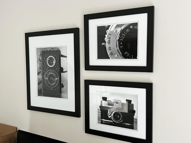 K&ouml;nnte beinhalten: Drei gerahmte Schwarz-Wei&szlig;-Fotografien von Vintage-Kameras und Kamerateilen. Die Bilder werden in schwarzen Rahmen mit wei&szlig;en Passepartouts pr&auml;sentiert. Die Fotografien zeigen Nahaufnahmen von Kameraobjektiven und dem Geh&auml;use einer Vintage-Kamera.