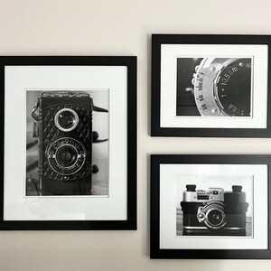 K&ouml;nnte beinhalten: Drei gerahmte Schwarz-Wei&szlig;-Fotografien von Vintage-Kameras. Die Bilder zeigen eine Nahaufnahme eines Kameraobjektivs, eine klassische Boxkamera und eine Kompaktkamera, alle mit wei&szlig;em Passepartout.
