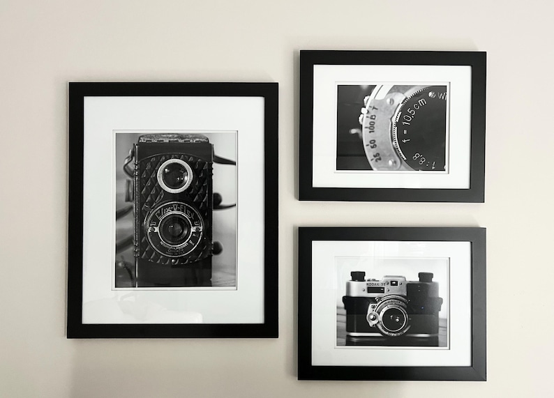 K&ouml;nnte beinhalten: Drei gerahmte Schwarz-Wei&szlig;-Fotografien von Vintage-Kameras. Die Bilder zeigen eine Nahaufnahme eines Kameraobjektivs, eine klassische Boxkamera und eine Kompaktkamera, alle mit wei&szlig;em Passepartout.
