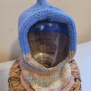 May include: A knitted hood in shades of blue, tan, and pastel stripes. The hood has a pointed top and a cowl neck. The item is made of a soft, textured yarn and is likely handmade. The hood is displayed on a woven basket.