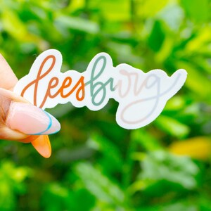 May include: A white sticker with the word "Leesburg" in a script font. The letters are in shades of orange, green, and pink. The sticker is held against a background of green foliage.