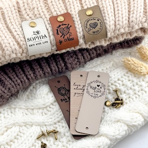 May include: A collection of rectangular faux leather labels in white, tan, and brown, featuring custom text and graphics like "Sophia Knit With Love" and "Handmade With Love." The labels include brass-colored metal rivets for attachment to knit items.