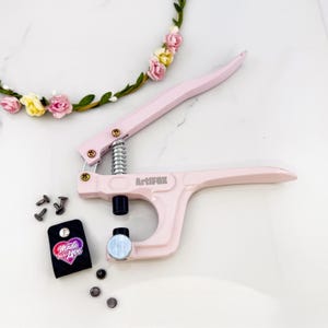 May include: A pink Artifox hand press tool with a silver spring and a black base. The tool is next to a black tag with the words "Made with Love" and a heart design. Several silver rivets and a floral crown are also in the image.