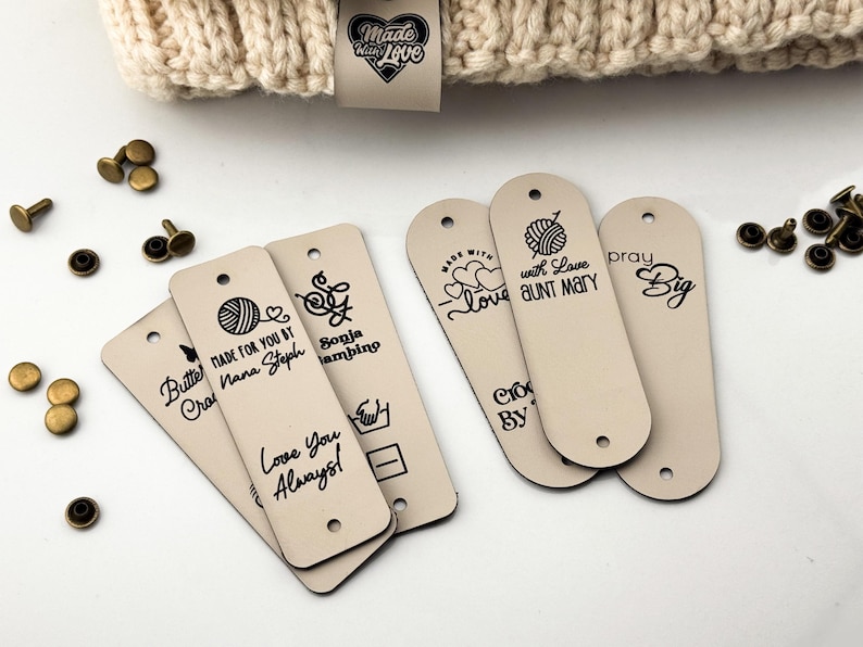 Puede incluir: Etiquetas rectangulares de piel sint&eacute;tica beige con extremos redondeados, con texto e iconos impresos en negro que incluyen ovillos de lana, corazones y nombres como Nana Steph y Aunt Mary. Incluye remaches de metal bronce.