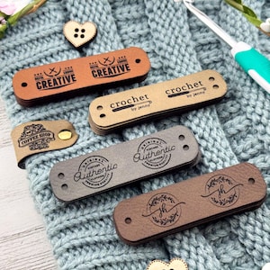 May include: A collection of rectangular faux leather clothing labels in various shades of brown and gray, featuring stamped designs including text like Creative, crochet by jenny, Coffee Shop, and Authentic, alongside a heart-shaped button and a crochet hook.