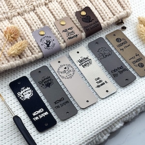May include: A collection of rectangular faux leather labels in black, gray, beige, and brown, featuring engraved text and graphics like hearts and yarn balls. Some labels are folded with brass rivets, while others are flat with pre-punched holes.