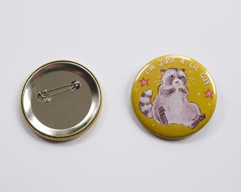May include: A yellow button with a raccoon illustration and the text "I'm just a lil guy". The raccoon is wearing a surprised expression and has a pink star on either side of it. The button has a gold border and a silver pin back.