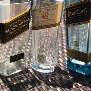 May include: Three empty glass bottles with labels for Johnnie Walker Black Label, Johnnie Walker 18 year old blended Scotch whisky, and Johnnie Walker Blue Label blended Scotch whisky. The bottles are sitting on a woven wicker surface.