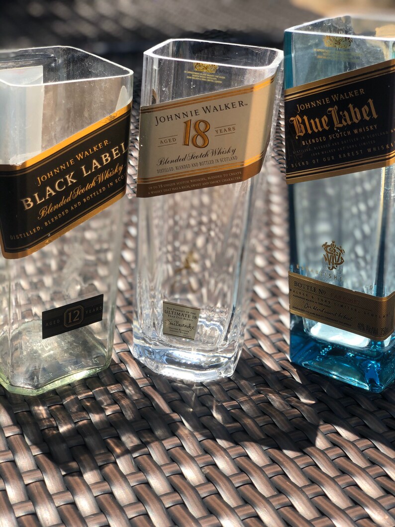 May include: Three empty glass bottles with labels for Johnnie Walker Black Label, Johnnie Walker 18 year old blended Scotch whisky, and Johnnie Walker Blue Label blended Scotch whisky. The bottles are sitting on a woven wicker surface.