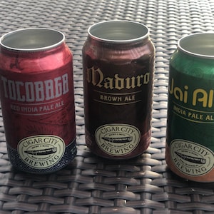 May include: Three aluminum cans of craft beer from Cigar City Brewing. The cans are labeled "Tocobaga Red India Pale Ale", "Maduro Brown Ale", and "Jai Alai India Pale Ale".