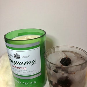 K&ouml;nnte beinhalten: Ein gr&uuml;nes Glas-Kerzengef&auml;&szlig; mit einem wei&szlig;en Etikett, auf dem "Tanqueray Imported" und "London Dry Gin" steht. Die Kerze ist angez&uuml;ndet und das Gef&auml;&szlig; steht auf einer wei&szlig;en, flauschigen Oberfl&auml;che. Neben der Kerze steht ein Glas mit Eis und Brombeeren.