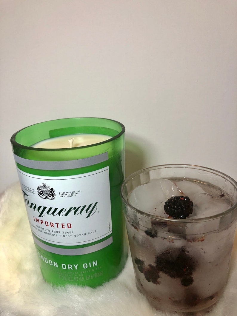 K&ouml;nnte beinhalten: Ein gr&uuml;nes Glas-Kerzengef&auml;&szlig; mit einem wei&szlig;en Etikett, auf dem "Tanqueray Imported" und "London Dry Gin" steht. Die Kerze ist angez&uuml;ndet und das Gef&auml;&szlig; steht auf einer wei&szlig;en, flauschigen Oberfl&auml;che. Neben der Kerze steht ein Glas mit Eis und Brombeeren.